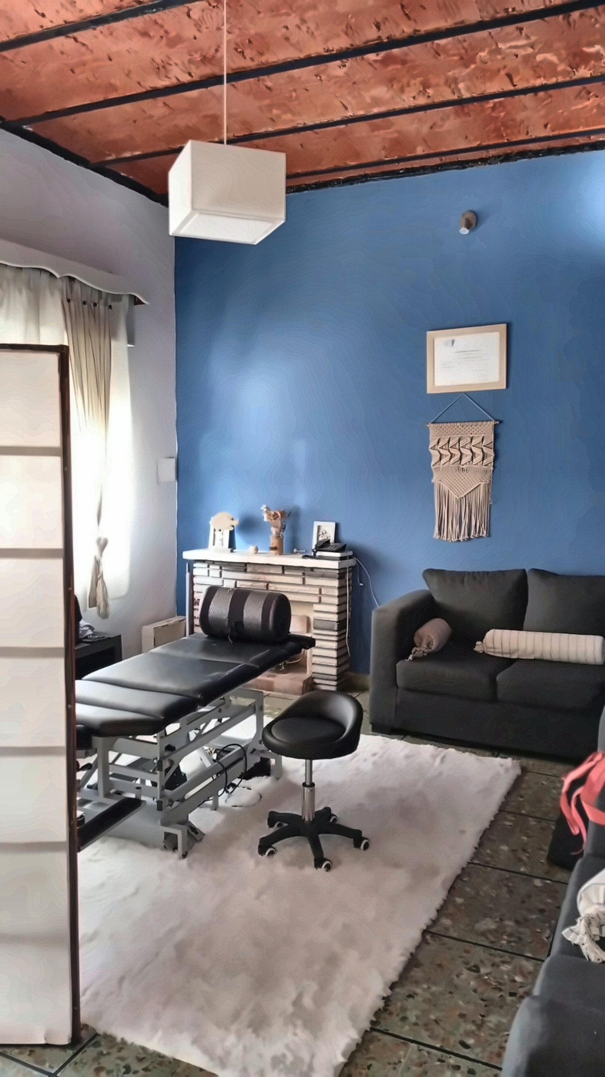 Sala del consultorio con pared azul, camilla negra sobre una alfombra blanca, banqueta, sillón gris y techo de ladrillo